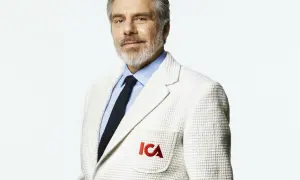 Man med grått skägg och blå skjorta, bär en vit kavaj med ICA-logga. Han ser ut att vara en del av nya ICA Stig.