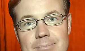 Morgan Olofsson, en man med glasögon och rött hår, ler mot kameran. Bakgrunden är orange.