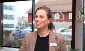 Kreativa Karolina, med ett leende och en brun tweedkavaj, deltar i ett möte. Bakom henne syns en stadsmiljö genom ett fönster.