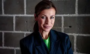 Amanda Schulman ung, med blå ögon och en grön tröja under en randig kavaj, ler mot kameran framför en tegelvägg.