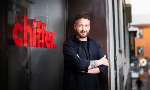 Gary Engman, med tatueringar och skägg, står med armarna i kors utanför en lokal med röd text som lyder "chiffer".