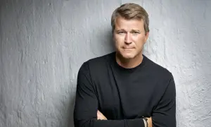 Anders Jensen, en man med blont hår och blå ögon, bär en svart tröja och står med armarna i kors mot en vit, strukturerad vägg.