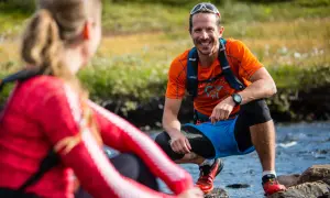 Två löpare i gorp core-stil vid en bäck. Mannen ler glatt i orange tröja och blå shorts.