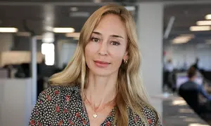 A blonde woman with a floral shirt and a heart necklace smiles at the camera in an office setting.