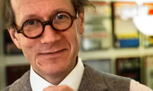 Johan Hakelius, en man med runda glasögon, ler mot kameran. Bakom honom syns bokhyllor.