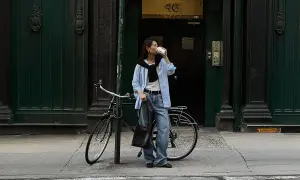 En kvinna i avslappnad street style dricker kaffe bredvid sin cykel framför en mörkgrön dörr.