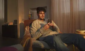 Ung man sitter i en fåtölj och tittar på sin mobil. En skål med mat står på ett bord bredvid. Kanske en paus från tre reklam.