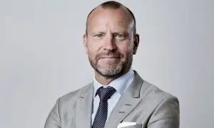 Peter Fellman, en man med gråsprängt skägg och blå ögon, bär en ljusgrå kavaj och randig slips.
