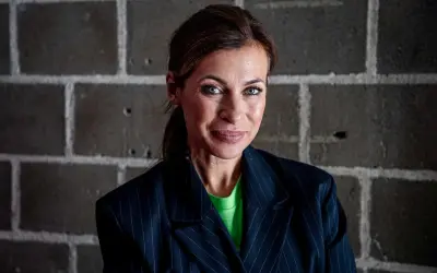 Amanda Schulman ung, med blå ögon och en grön tröja under en randig kavaj, ler mot kameran framför en tegelvägg.