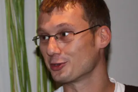 Sladjan Osmanagic, en man med glasögon, ler mot kameran. Bakom honom syns gröna växter.