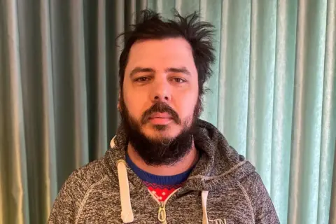 Man med rufsigt hår och skägg, klädd i en grå hoodie över en julig tröja. Han ser ut att ha haft en chang frick.