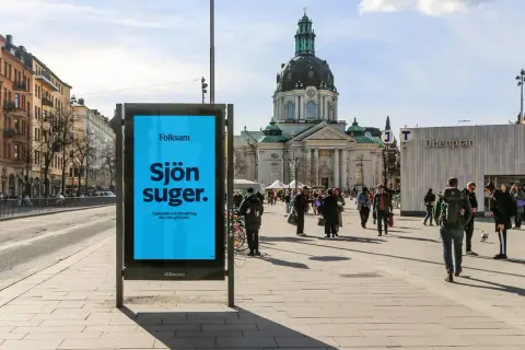 Folksam reklam på en blå skylt i en stadsmiljö. "Sjön suger" står det, med texten "Sparande och försäkring ska inte göra det." under.