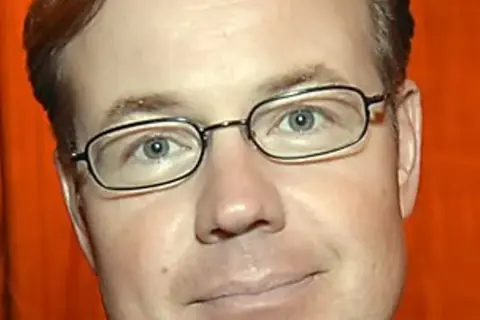 Morgan Olofsson, en man med glasögon och rött hår, ler mot kameran. Bakgrunden är orange.
