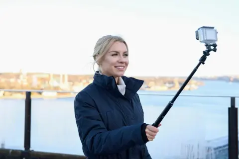 Therese Lindgren filmar med GoPro på selfiepinne vid en vattensamling med stad i bakgrunden.