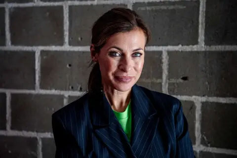 Amanda Schulman ung, med blå ögon och en grön tröja under en randig kavaj, ler mot kameran framför en tegelvägg.