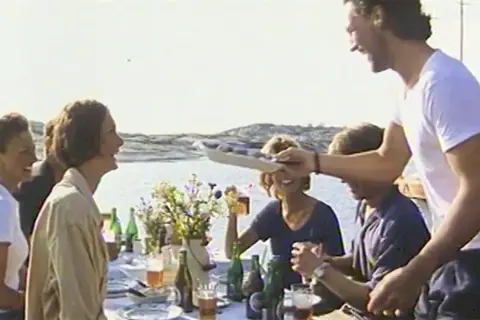 En glad grupp njuter av en sommarmåltid vid havet, en scen som påminner om en klassisk pripps blå reklam.