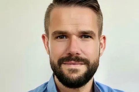 Johnny Kroneld, en man med kortklippt mörkt hår och skägg, bär en blå skjorta.