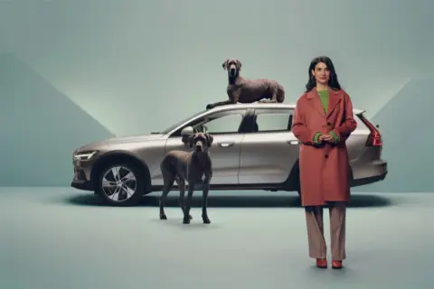 En kvinna och två stora hundar står bredvid en silverfärgad Volvo. En hund vilar på taket, den andra står framför bilen. Volvo reklam.