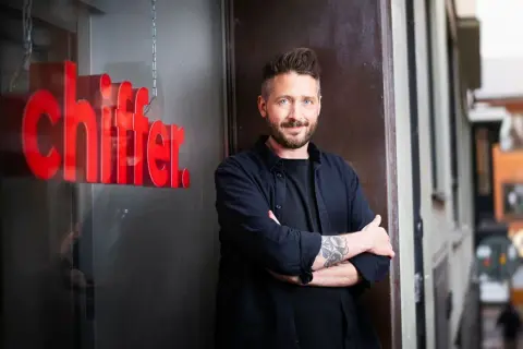 Gary Engman, med tatueringar och skägg, står med armarna i kors utanför en lokal med röd text som lyder "chiffer".