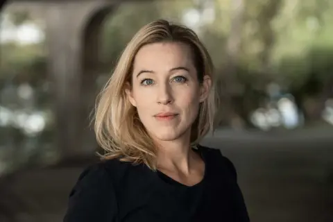 Anna Careborg, en kvinna med blont hår och blå ögon, tittar rakt fram. Bakgrunden är suddig med grönska och en valvformad struktur.