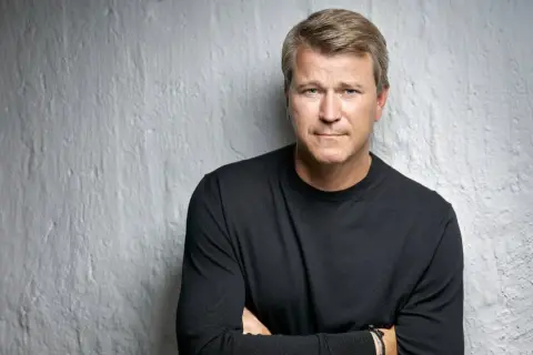 Anders Jensen, en man med blont hår och blå ögon, bär en svart tröja och står med armarna i kors mot en vit, strukturerad vägg.