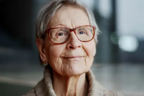 Kerstin Brunnberg, en äldre kvinna med glasögon och grått hår, ler mjukt.