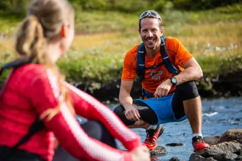Två löpare i gorp core-stil vid en bäck. Mannen ler glatt i orange tröja och blå shorts.