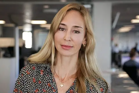 A blonde woman with a floral shirt and a heart necklace smiles at the camera in an office setting.