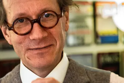 Johan Hakelius, en man med runda glasögon, ler mot kameran. Bakom honom syns bokhyllor.