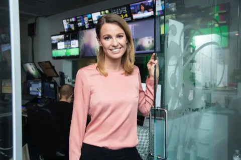Jennie Pierrou ler vid en glasdörr i en kontrollrumsmiljö med många skärmar.