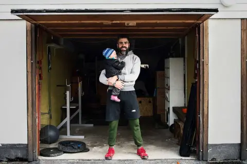 Man holding a baby in a garage with gym equipment.