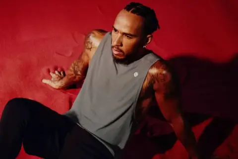 Lewis Hamilton visar upp sin Lewis Hamilton fashion-stil i en grå linne och svarta byxor, sittandes på en röd yta.