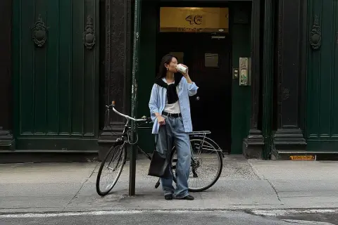 En kvinna i avslappnad street style dricker kaffe bredvid sin cykel framför en mörkgrön dörr.