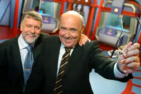 Ingvar Oldsberg, med ett brett leende, håller upp sina glasögon. Han står bredvid en annan man i en studio.