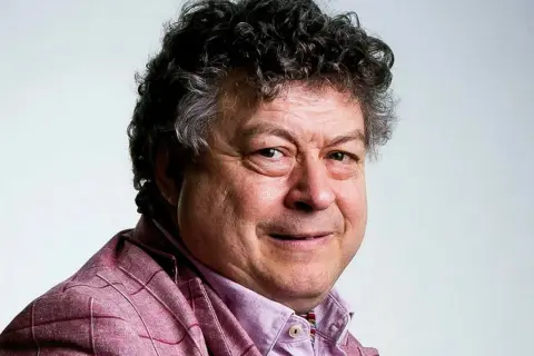 Rory Sutherland, med sitt karaktäristiska lockiga hår och ett vänligt leende, bär en rutig kavaj.
