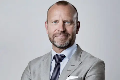 Peter Fellman, en man med gråsprängt skägg och blå ögon, bär en ljusgrå kavaj och randig slips.