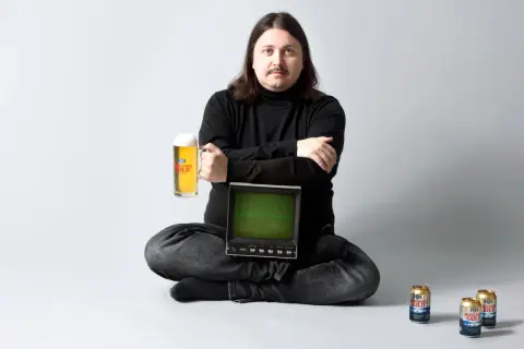 Man med mustasch sitter med en öl i handen och en gammal datorskärm. Norrlands Guld reklam.