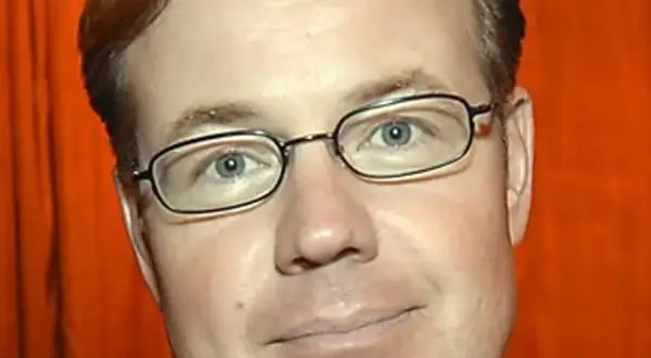 Morgan Olofsson, en man med glasögon och rött hår, ler mot kameran. Bakgrunden är orange.