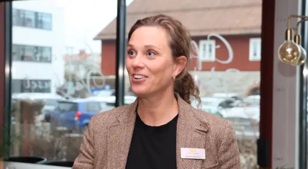 Kreativa Karolina, med ett leende och en brun tweedkavaj, deltar i ett möte. Bakom henne syns en stadsmiljö genom ett fönster.