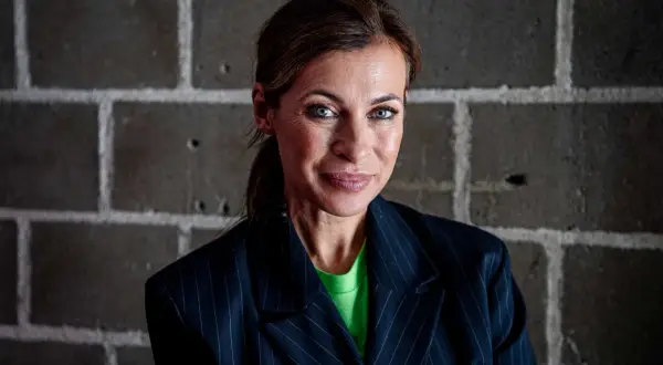 Amanda Schulman ung, med blå ögon och en grön tröja under en randig kavaj, ler mot kameran framför en tegelvägg.