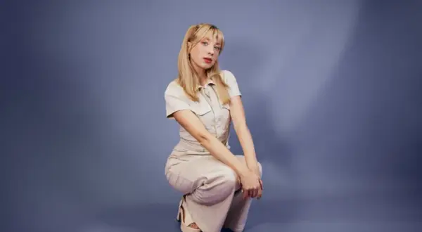 Sandra Beijer sitter på huk i en ljusbeige dräkt mot en blå bakgrund.