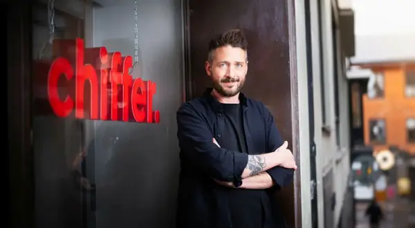 Gary Engman, med tatueringar och skägg, står med armarna i kors utanför en lokal med röd text som lyder "chiffer".