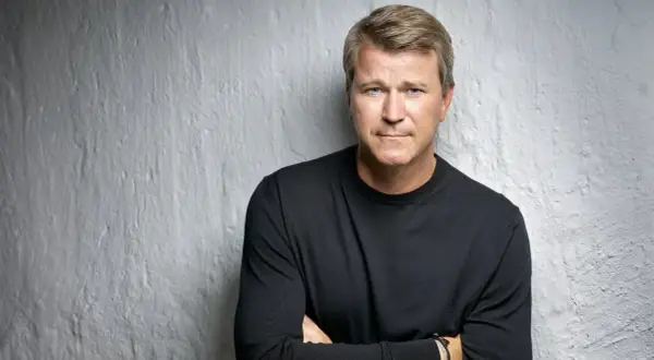 Anders Jensen, en man med blont hår och blå ögon, bär en svart tröja och står med armarna i kors mot en vit, strukturerad vägg.