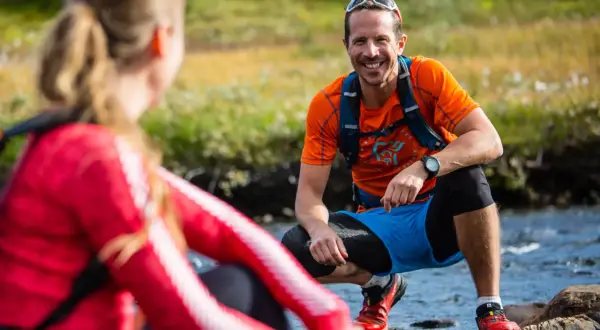 Två löpare i gorp core-stil vid en bäck. Mannen ler glatt i orange tröja och blå shorts.