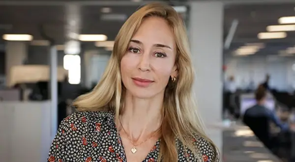 A blonde woman with a floral shirt and a heart necklace smiles at the camera in an office setting.