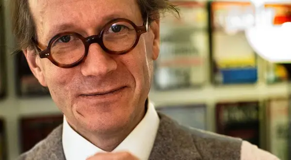 Johan Hakelius, en man med runda glasögon, ler mot kameran. Bakom honom syns bokhyllor.