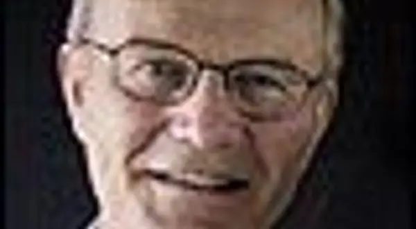 Närbild på Olle Stenholm med glasögon, ett vänligt leende och mörkt bakgrund.