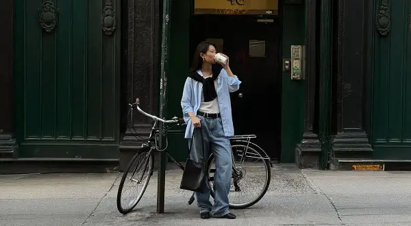 En kvinna i avslappnad street style dricker kaffe bredvid sin cykel framför en mörkgrön dörr.
