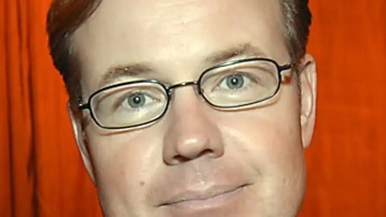 Morgan Olofsson, en man med glasögon och rött hår, ler mot kameran. Bakgrunden är orange.