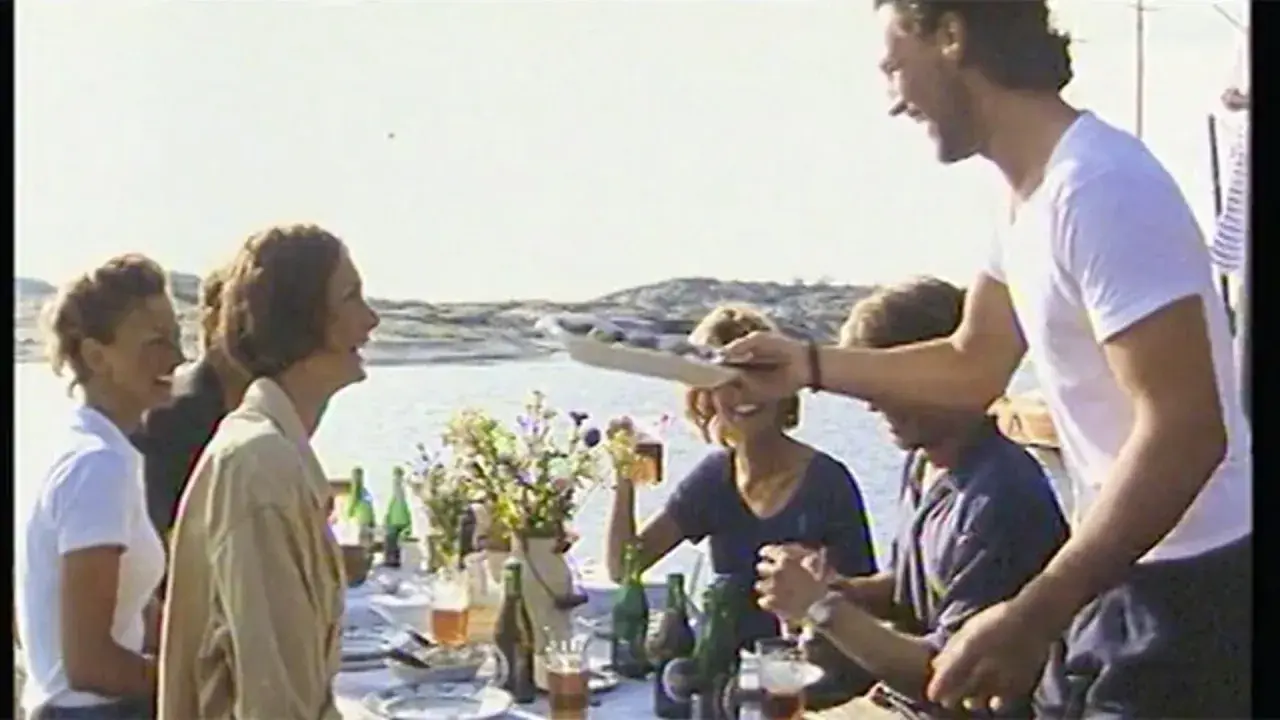 En glad grupp njuter av en sommarmåltid vid havet, en scen som påminner om en klassisk pripps blå reklam.